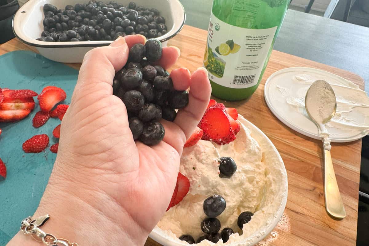 A hand holding fresh blueberries over a bowl of whipped cream topped with strawberries and blueberries, with a cutting board, sliced strawberries, a colander of blueberries, and a bottle of lemon juice in the background.