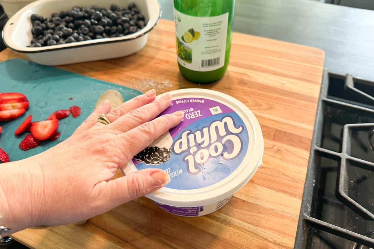A hand rests on a container of Cool Whip placed on a wooden countertop next to sliced strawberries, blueberries in a dish, a green bottle, and a kitchen stove.