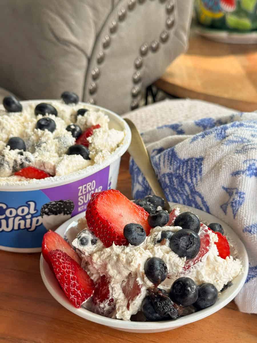 A bowl of Cool Whip Zero Sugar dessert topped with fresh strawberries and blueberries sits in front of its container. A spoon, a blue and white towel, and a cushioned chair are in the background.