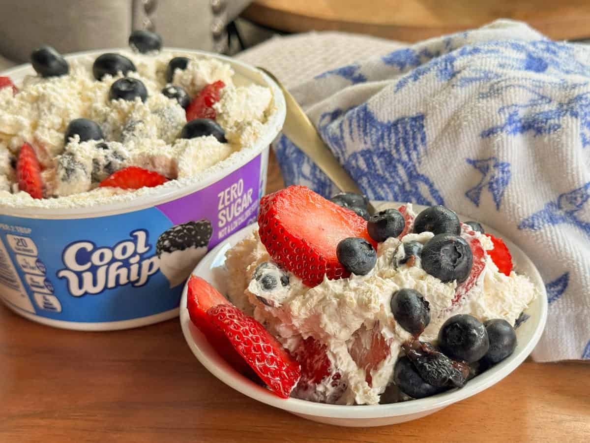 A bowl of Cool Whip Zero Sugar topped with fresh strawberries and blueberries sits in front of its container. A spoon and a blue-and-white patterned cloth are in the background.