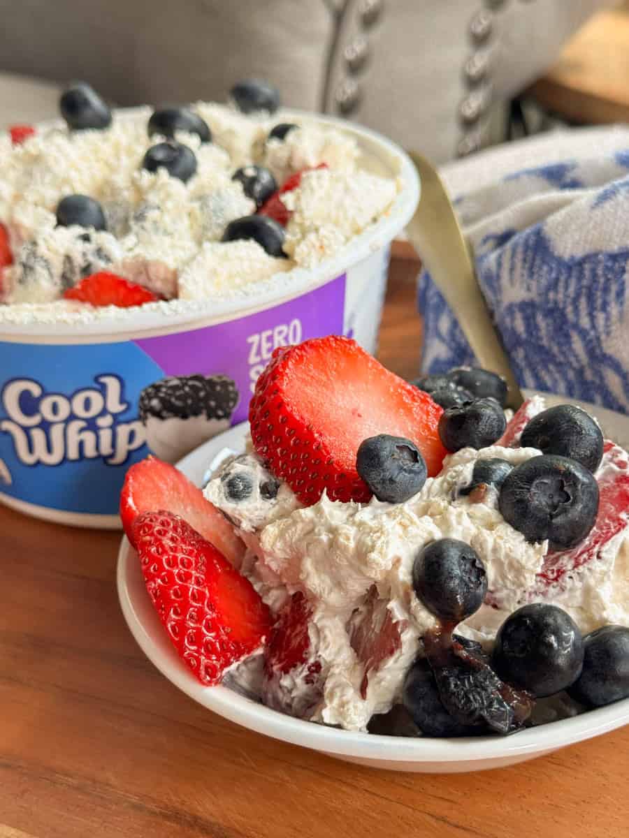 A bowl of whipped topping mixed with sliced strawberries and blueberries sits in front of a container of Cool Whip Zero. A spoon and a blue-and-white towel are nearby on a wooden surface.