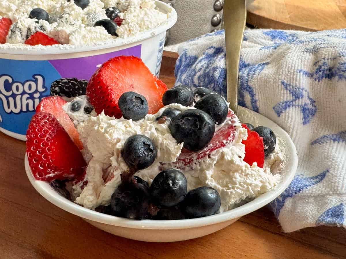 A small bowl filled with Cool Whip, topped with fresh strawberries and blueberries. A spoon rests in the bowl, and a container of Cool Whip and a blue-and-white towel are nearby on a wooden surface.