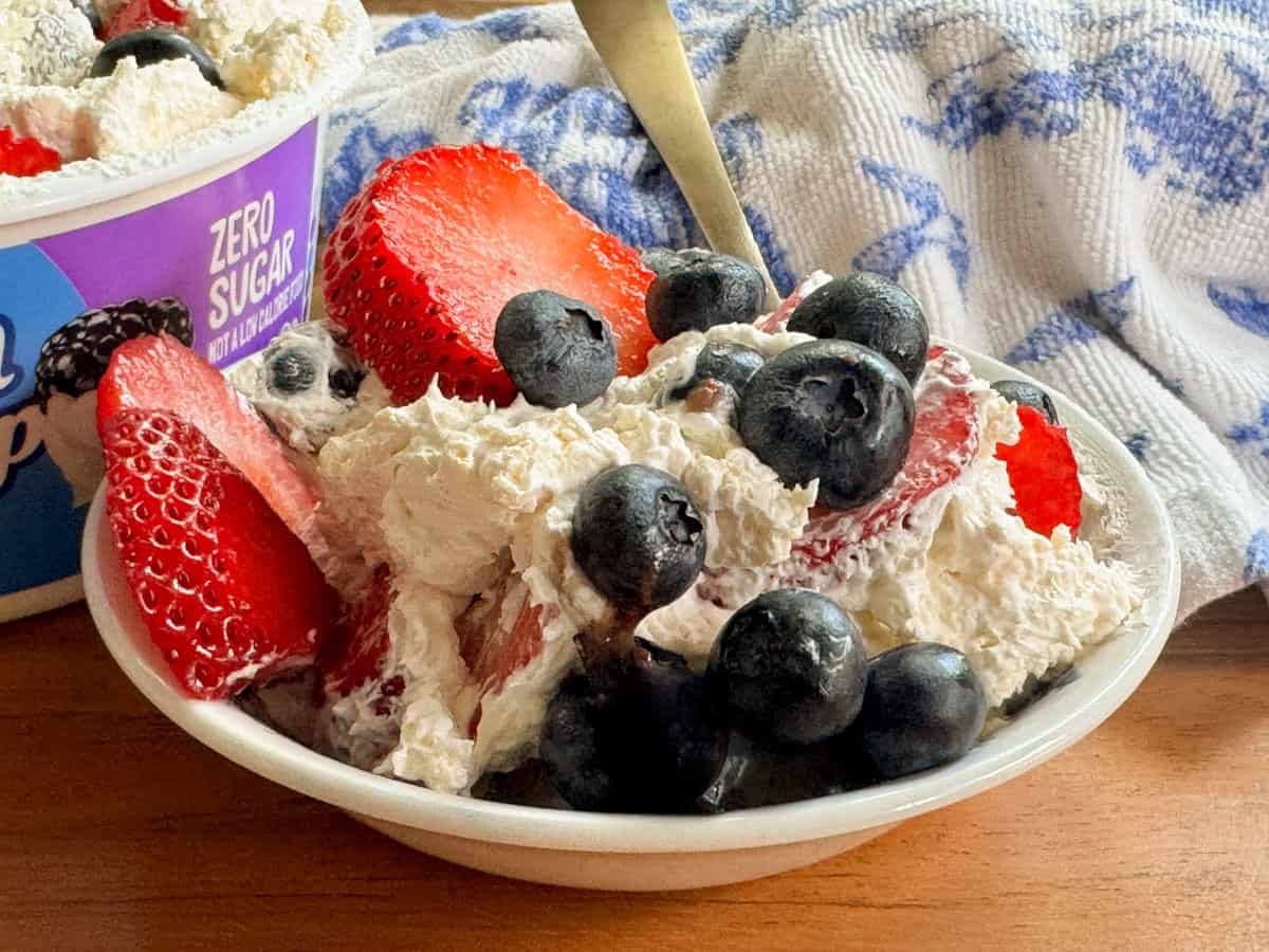 A small bowl filled with whipped cream, sliced strawberries, and blueberries sits on a wooden surface. A spoon and a container labeled "zero sugar" are visible in the background.