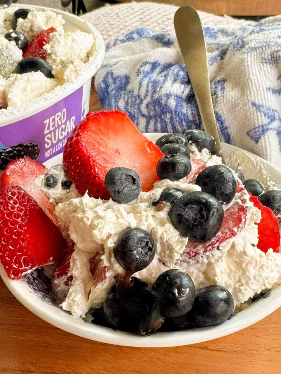 A bowl of whipped cream topped with sliced strawberries and whole blueberries sits next to a container labeled "Zero Sugar." A spoon rests in the bowl, and a blue and white cloth is in the background.