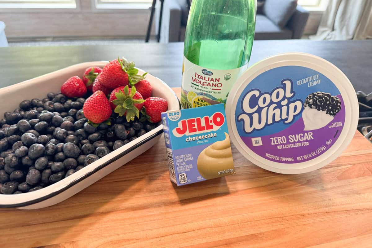 A white tray of blueberries and strawberries sits on a wooden counter next to a bottle of lemon juice, a container of Cool Whip Zero Sugar, and a box of Jell-O cheesecake pudding mix.